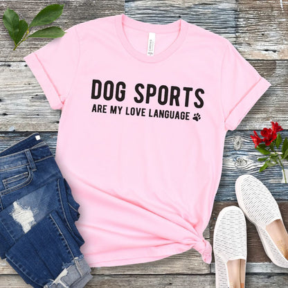 Pink t-shirt with 'Dog Sports are my love language' text on a wooden surface with jeans and shoes.