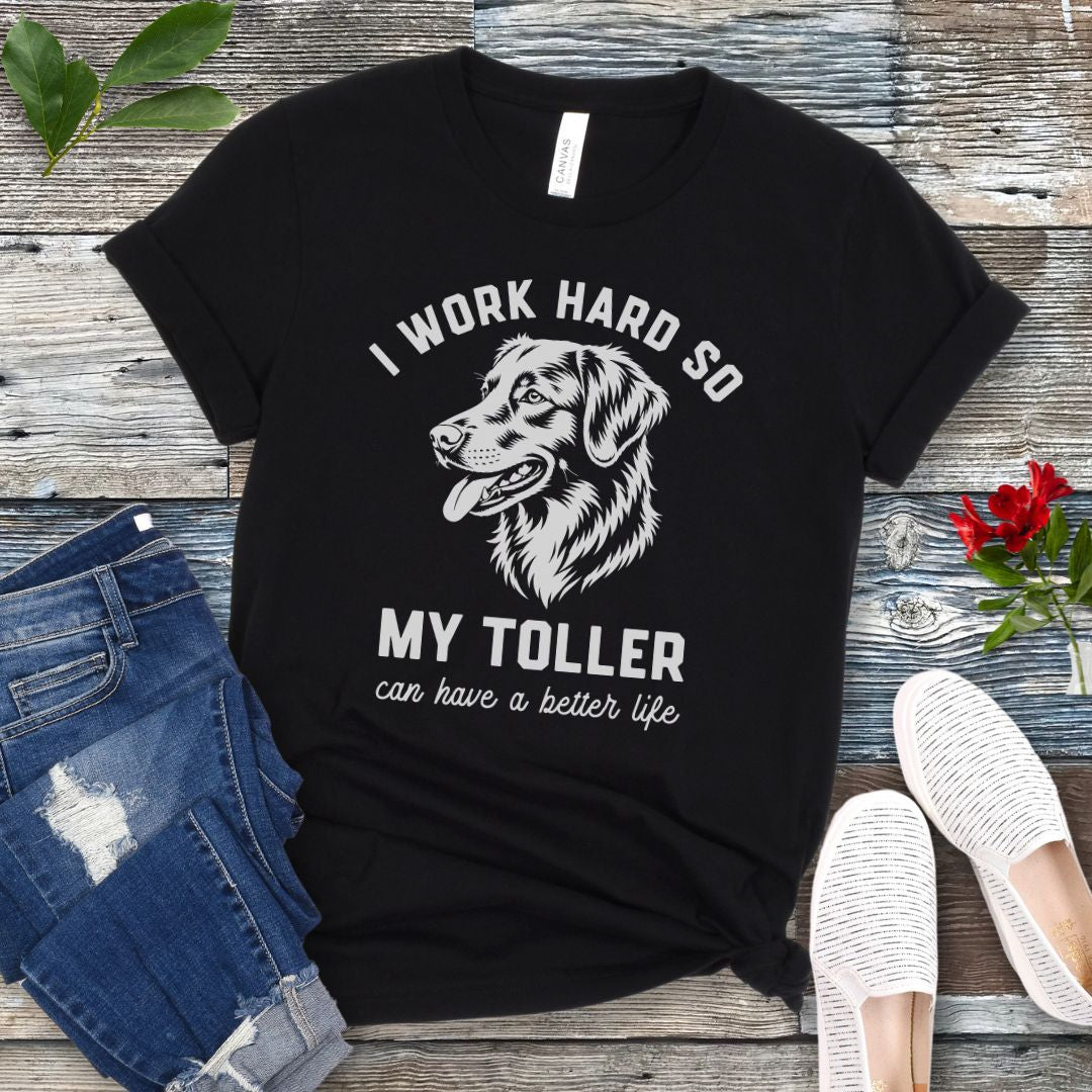 Black unisex t-shirt for Nova Scotia Duck Tolling Retriever lovers with linocut-style breed head study graphic and humorous text 'I work hard so my Toller can have a better life', shown on a wooden surface with jeans and shoes. 