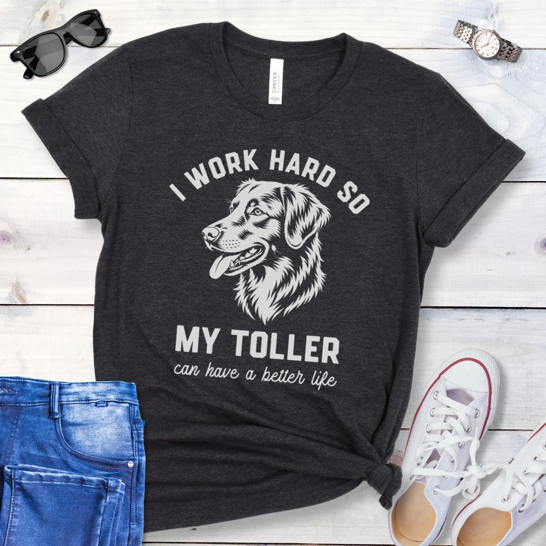 Dark grey unisex t-shirt for Nova Scotia Duck Tolling Retriever lovers with linocut-style breed head study graphic and humorous text 'I work hard so my Toller can have a better life', shown on a wooden surface with jeans and shoes. 