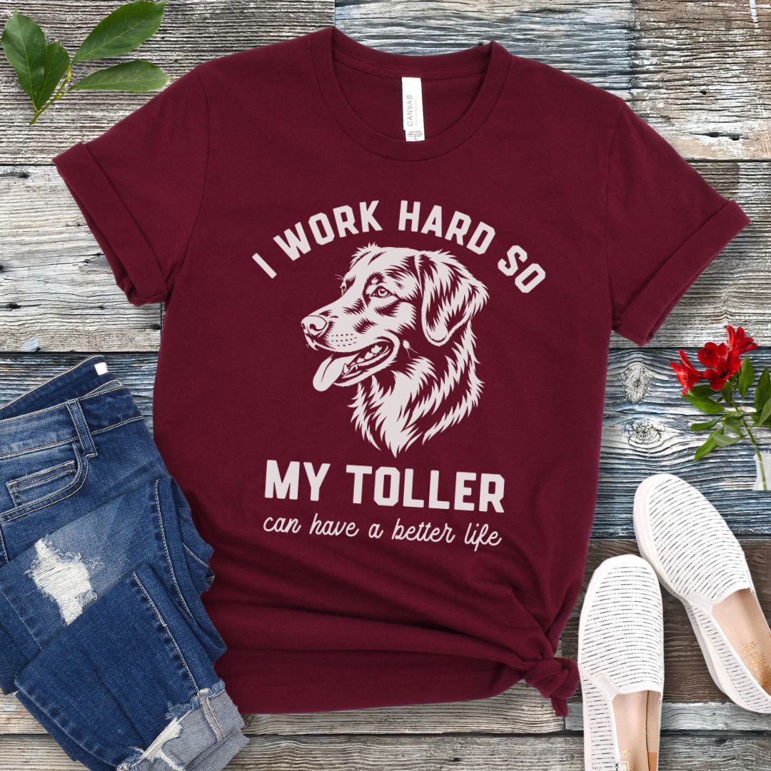 Maroon unisex t-shirt for Nova Scotia Duck Tolling Retriever lovers with linocut-style breed head study graphic and humorous text 'I work hard so my Toller can have a better life', shown on a wooden surface with jeans and shoes. 