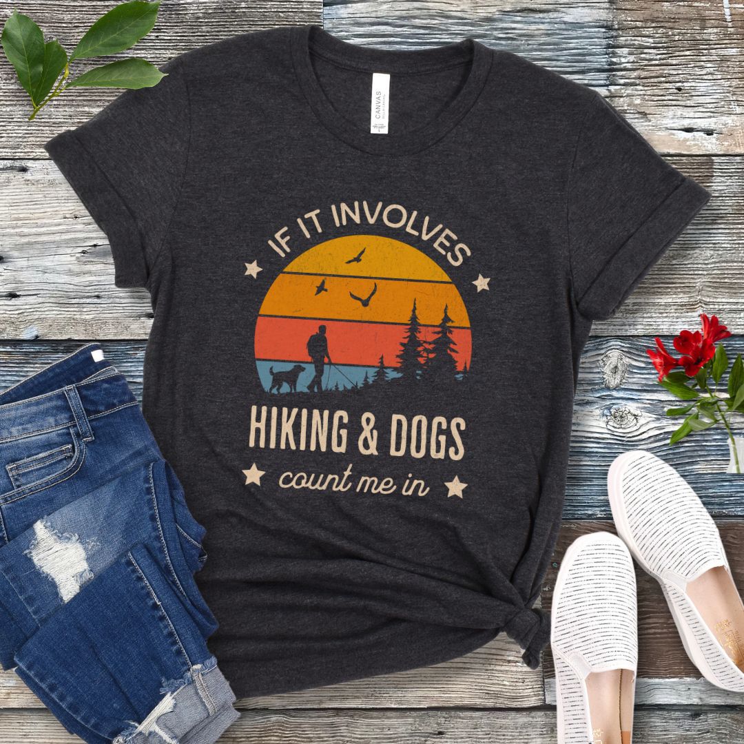 Dark grey T-shirt with text 'IF IT INVOLVES HIKING & DOGS COUNT ME IN' printed on the front, featuring a sunset scene and hiker and dog silhouettes. The shirt is laid out flat with a pair of blue jeans and white sneakers.