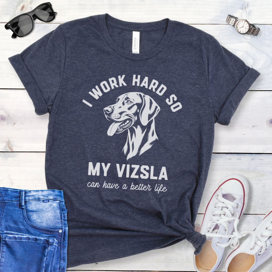 Heather navy blue unisex t-shirt for Hungarian Vizsla lovers with linocut-style Vizsla head study graphic and humorous text 'I work hard so my Vizsla can have a better life', shown on a wooden surface with jeans and shoes. 