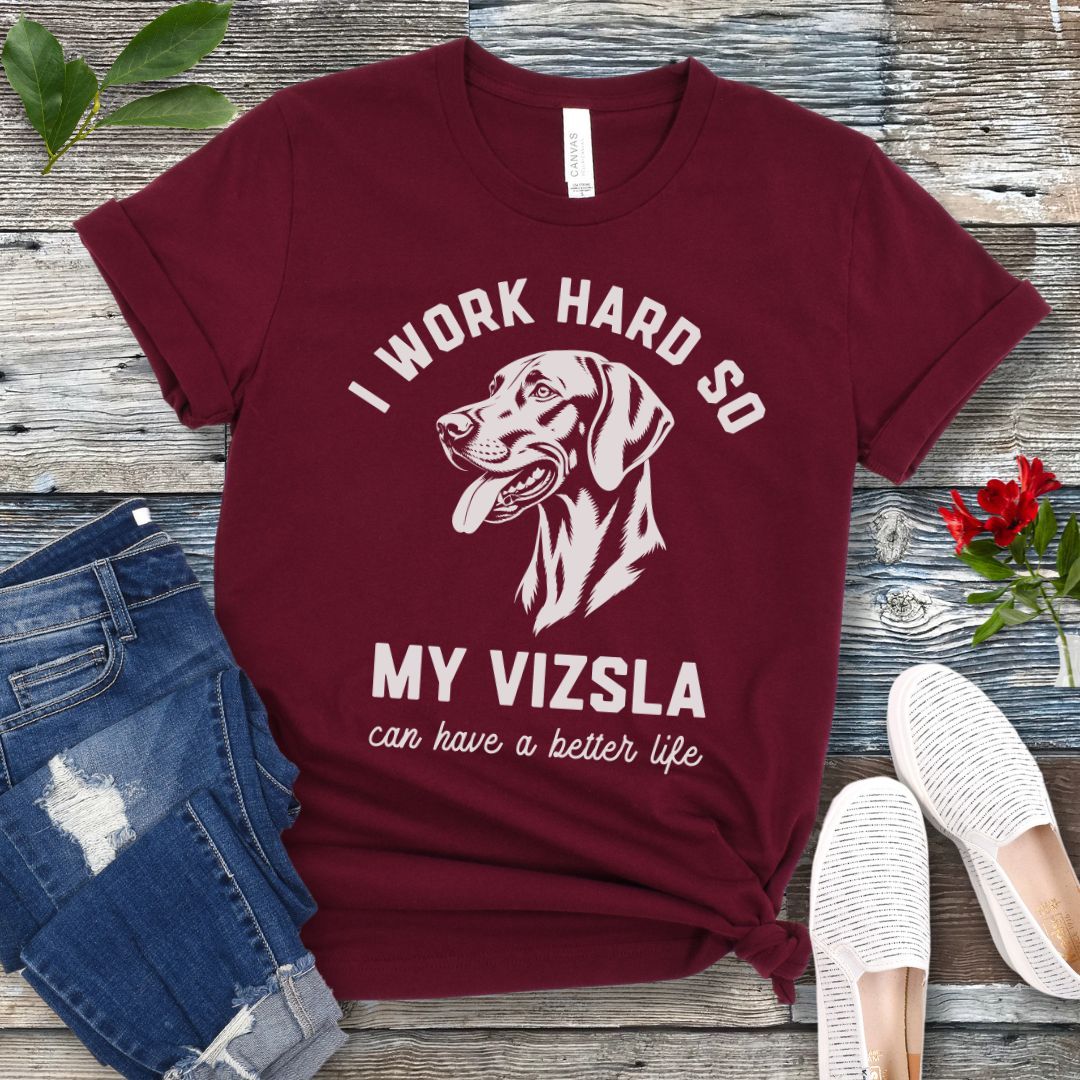 Maroon unisex t-shirt for Hungarian Vizsla lovers with linocut-style Vizsla head study graphic and humorous text 'I work hard so my Vizsla can have a better life', shown on a wooden surface with jeans and shoes. 