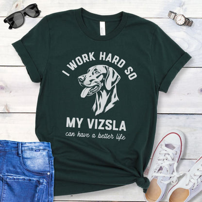 Dark green unisex t-shirt for Hungarian Vizsla lovers with linocut-style Vizsla head study graphic and humorous text 'I work hard so my Vizsla can have a better life', shown on a wooden surface with jeans and shoes. 