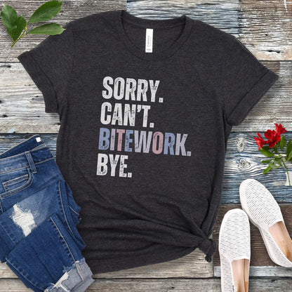 Dark grey protection dog sports t-shirt with colorful, distressed text 'SORRY. CAN'T. BITEWORK. BYE.', presented on a wooden surface with jeans and shoes.