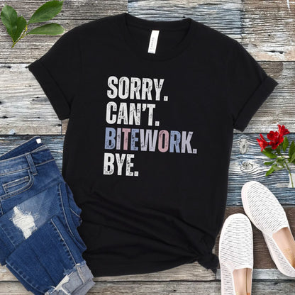 Black protection dog training t-shirt with colorful, distressed text 'SORRY. CAN'T. BITEWORK. BYE.', presented on a wooden surface with jeans and shoes.