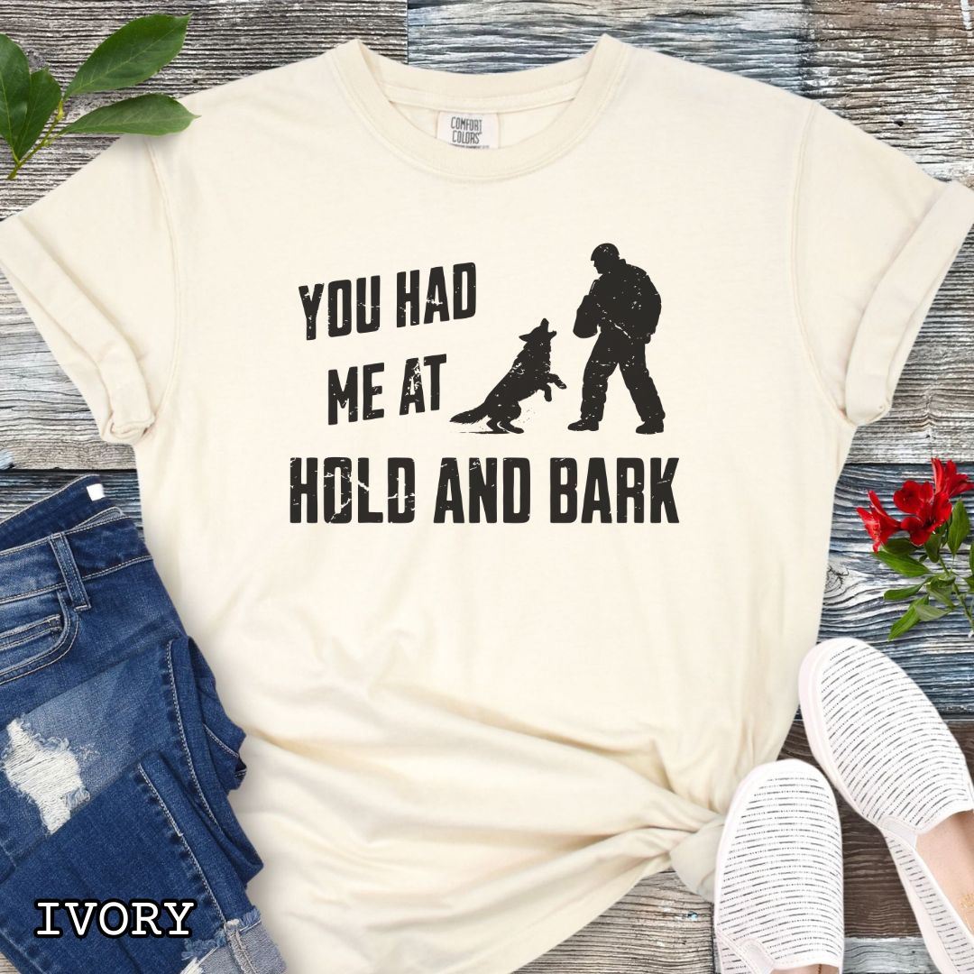 Ivory Comfort Colors t-shirt for protection dog sports handlers with graphic of a dog barking at a man in a bite suit and and text 'You had me at hold and bark', shown on a wooden surface.