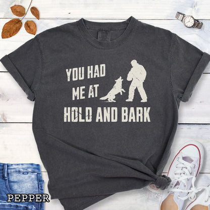 Dark grey Comfort Colors t-shirt for protection dog sports handlers with graphic of a dog barking at a man in a bite suit and and text 'You had me at hold and bark', shown on a wooden surface.