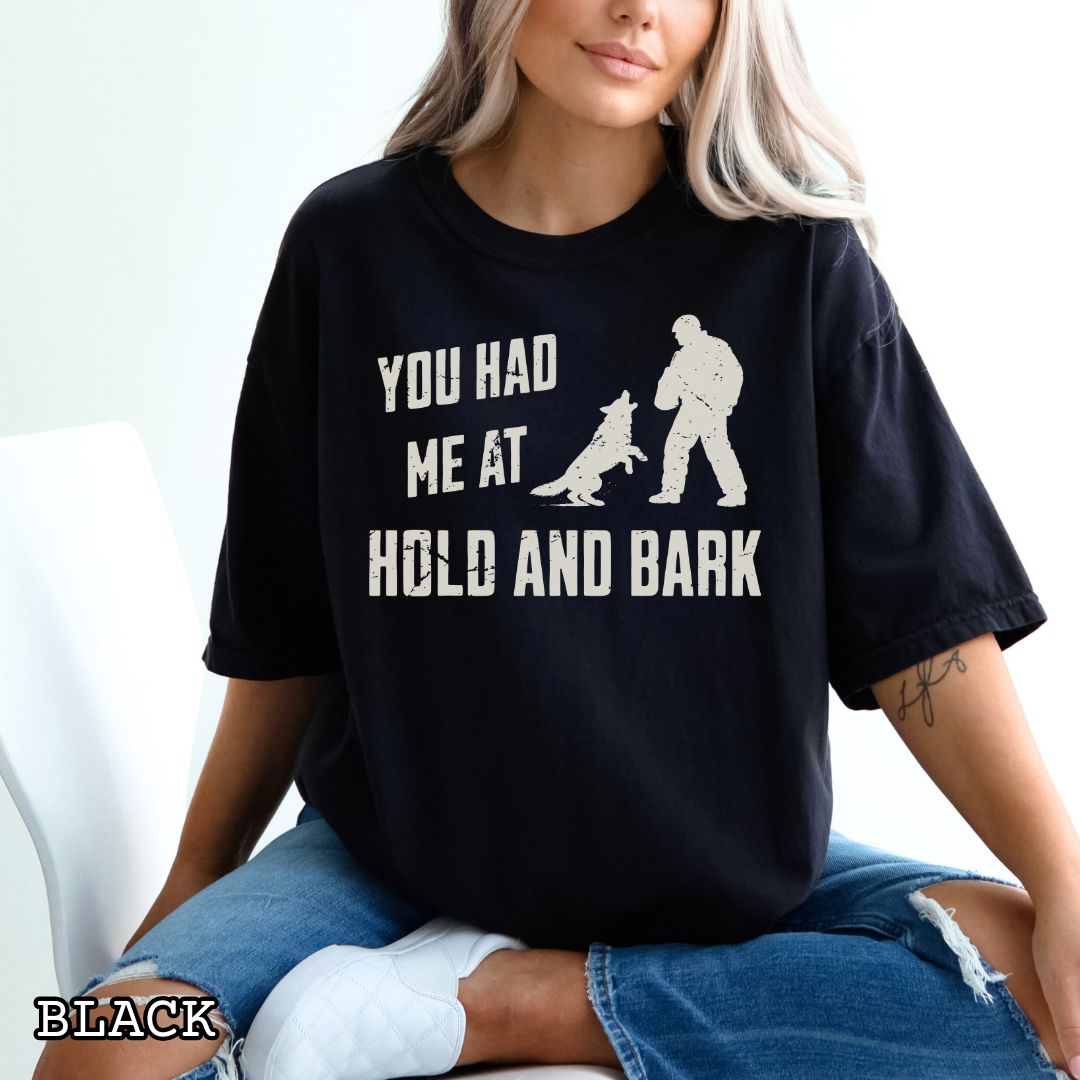 Black Comfort Colors t-shirt for protection dog sports handlers with graphic of a dog barking at a man in a bite suit and and text 'You had me at hold and bark', shown on a wooden surface.