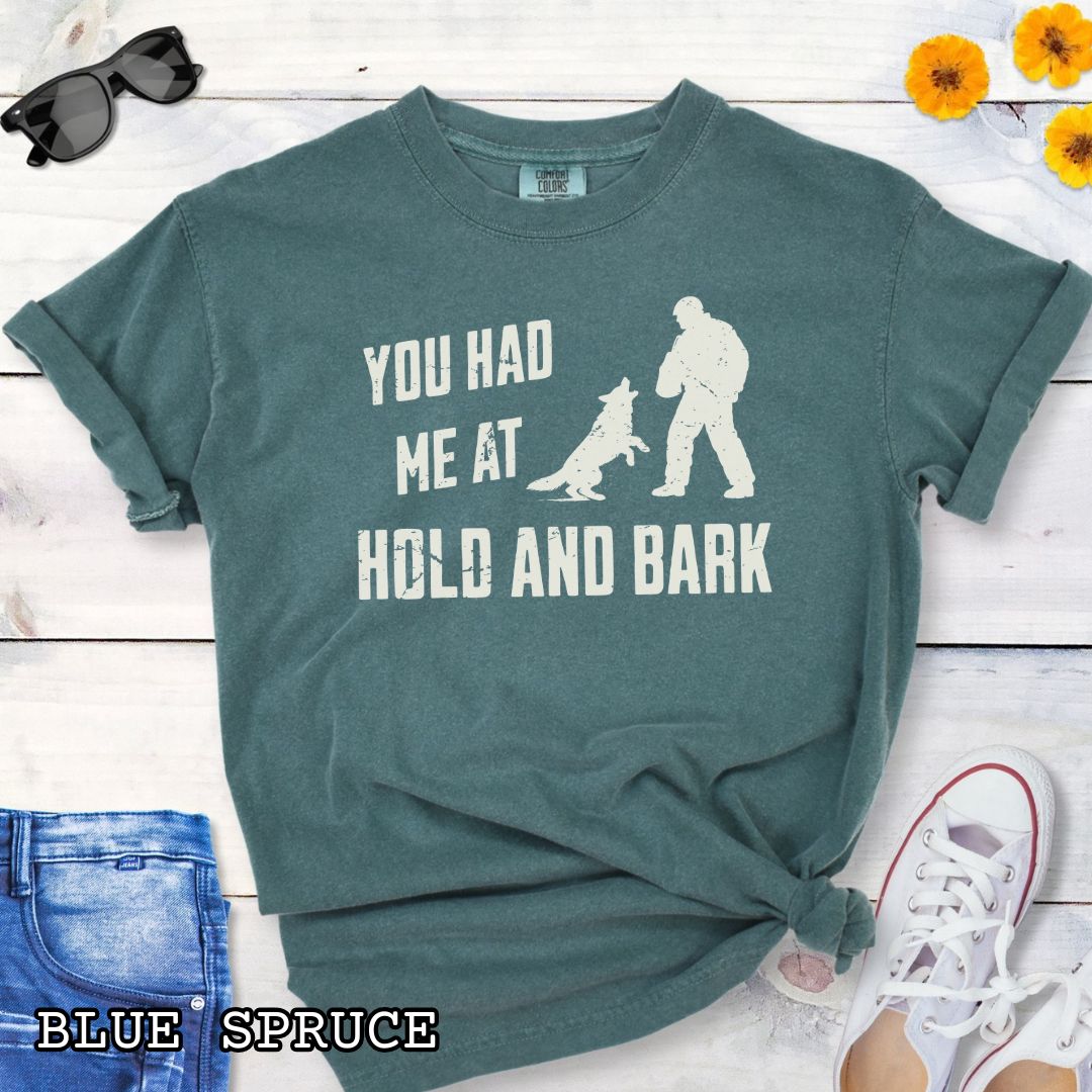 Green Comfort Colors t-shirt for protection dog sports handlers with graphic of a dog barking at a man in a bite suit and and text 'You had me at hold and bark', shown on a wooden surface.