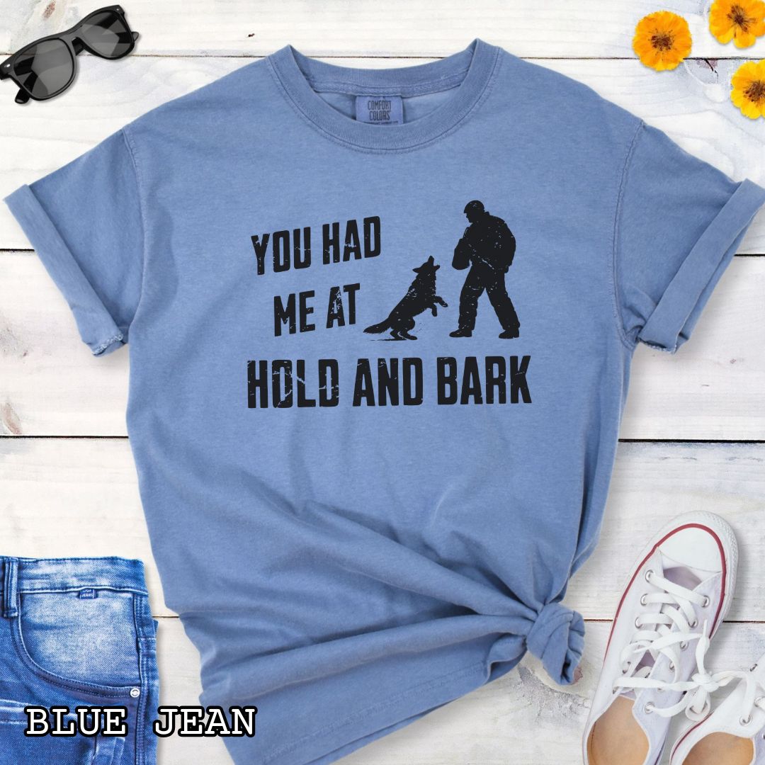 Blue Comfort Colors t-shirt for protection dog sports handlers with graphic of a dog barking at a man in a bite suit and and text 'You had me at hold and bark', shown on a wooden surface.