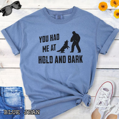 Blue Comfort Colors t-shirt for protection dog sports handlers with graphic of a dog barking at a man in a bite suit and and text 'You had me at hold and bark', shown on a wooden surface.
