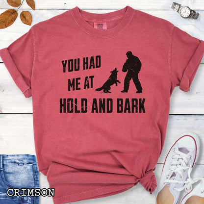 Red Comfort Colors t-shirt for protection dog sports handlers with graphic of a dog barking at a man in a bite suit and and text 'You had me at hold and bark', shown on a wooden surface.