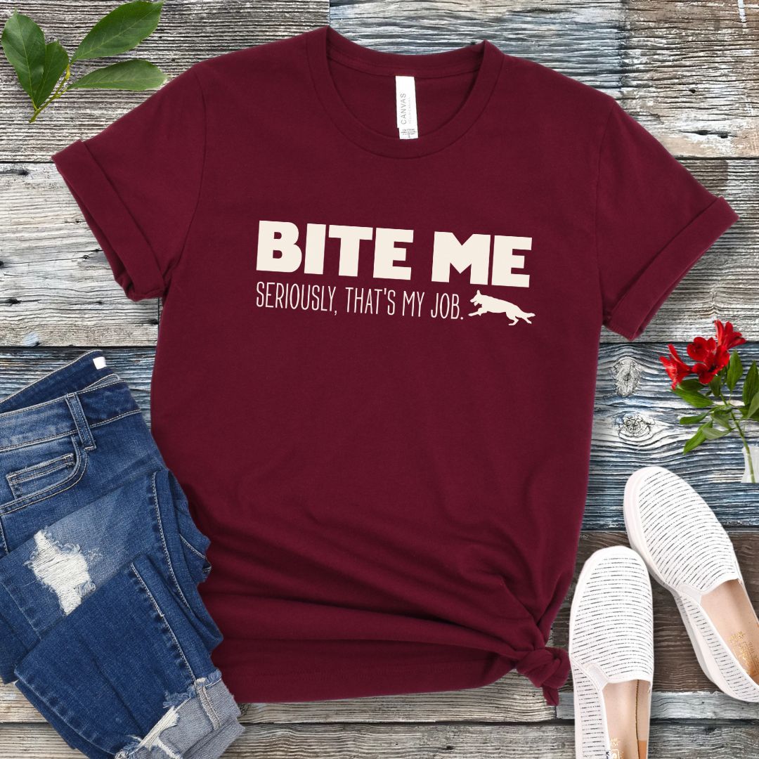 Maroon K9 helper t-shirt with 'BITE ME Seriously, That's My Job' text and running dog graphic, presented on a wooden surface with jeans and shoes.