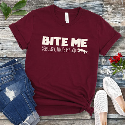 Maroon K9 helper t-shirt with 'BITE ME Seriously, That's My Job' text and running dog graphic, presented on a wooden surface with jeans and shoes.