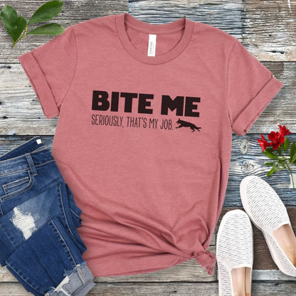 Mauve K9 helper t-shirt with 'BITE ME Seriously, That's My Job' text and running dog graphic, presented on a wooden surface with jeans and shoes.
