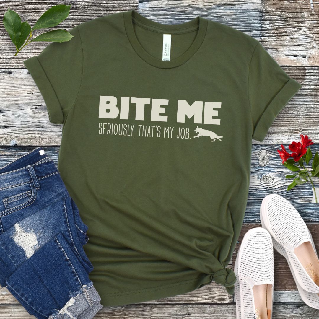 Military green K9 helper t-shirt with 'BITE ME Seriously, That's My Job' text and running dog graphic, presented on a wooden surface with jeans and shoes.