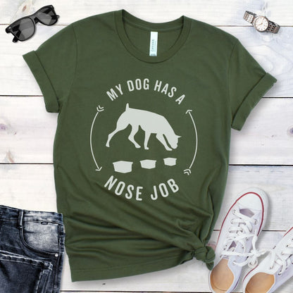 Military green dog canine scent work t-shirt with graphic of a searching dog with text 'MY DOG HAS A NOSE JOB' on the front, presented on a wooden surface with denim jeans and white shoes in the background.