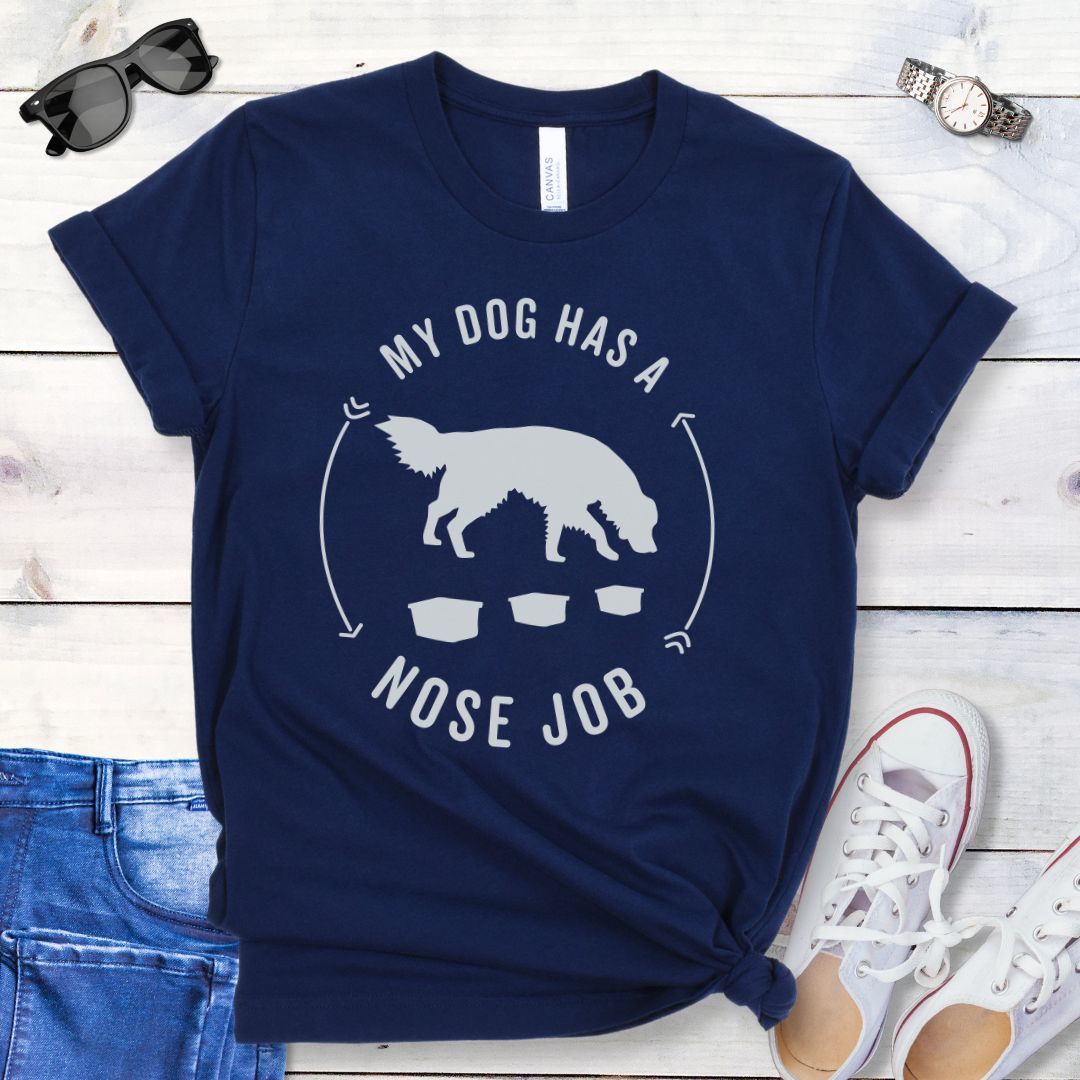 Navy blue dog nose work t-shirt with graphic of a searching dog with text 'MY DOG HAS A NOSE JOB' on the front, presented on a wooden surface with denim jeans and white shoes in the background.