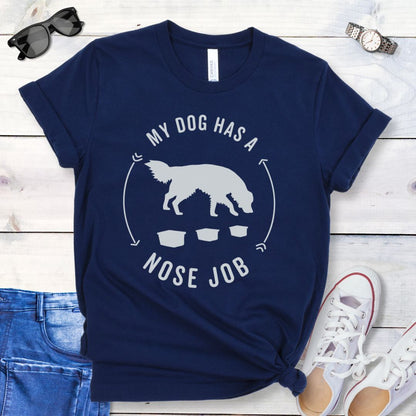 Navy blue dog nose work t-shirt with graphic of a searching dog with text 'MY DOG HAS A NOSE JOB' on the front, presented on a wooden surface with denim jeans and white shoes in the background.