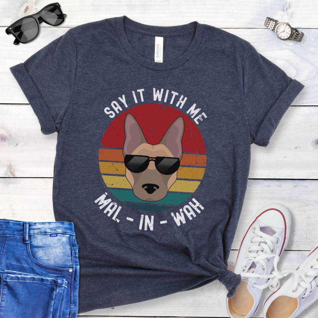 Heather navy t-shirt for Belgian Malinois lovers with a Malinois dog face wearing sunglasses, on a vintage sunset graphic, and humorous text 'Say it with me - Mal-in-wah', shown on a wooden surface with accessories.