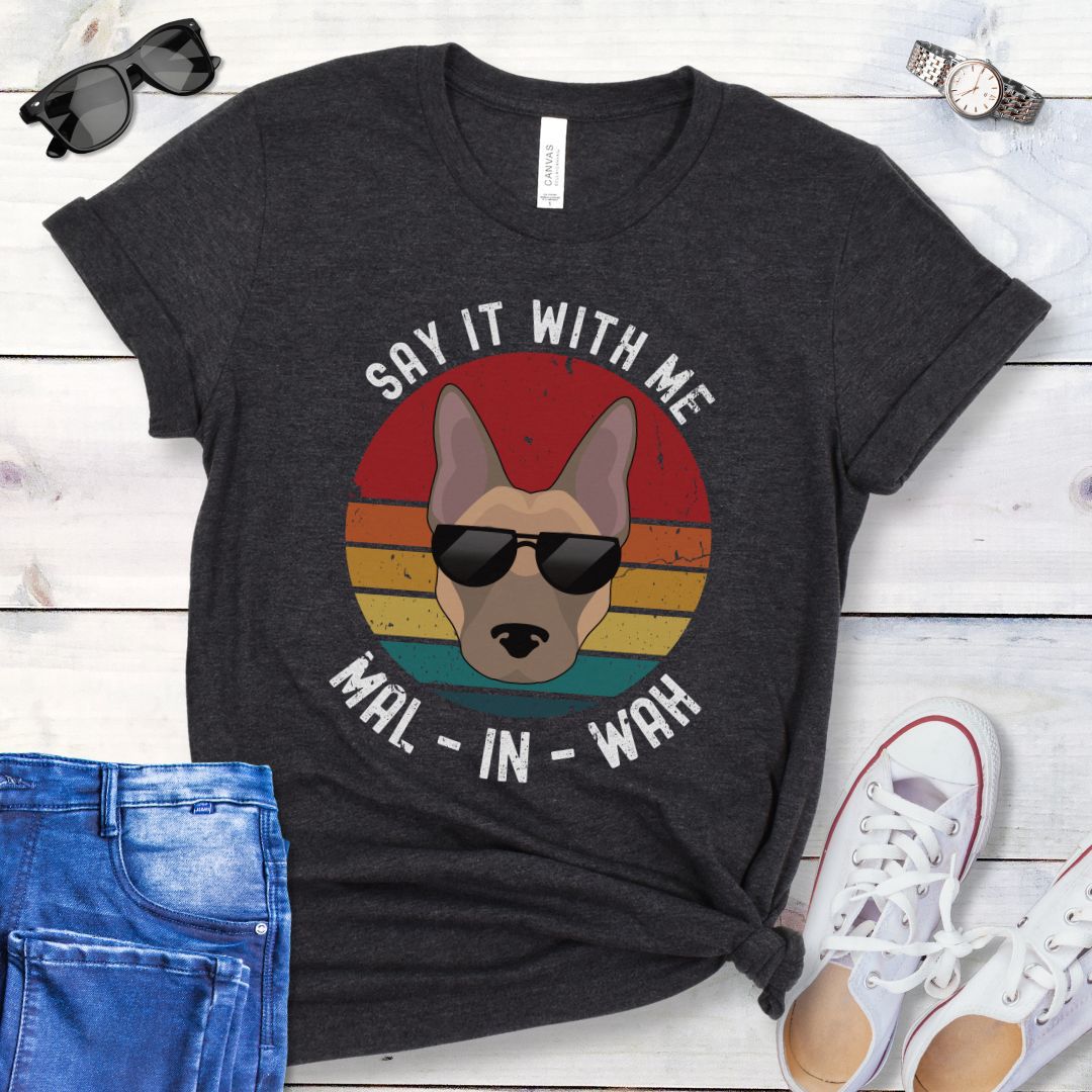 Dark grey t-shirt for Belgian Malinois lovers with a Malinois dog face wearing sunglasses, on a vintage sunset graphic, and humorous text 'Say it with me - Mal-in-wah', shown on a wooden surface with accessories.