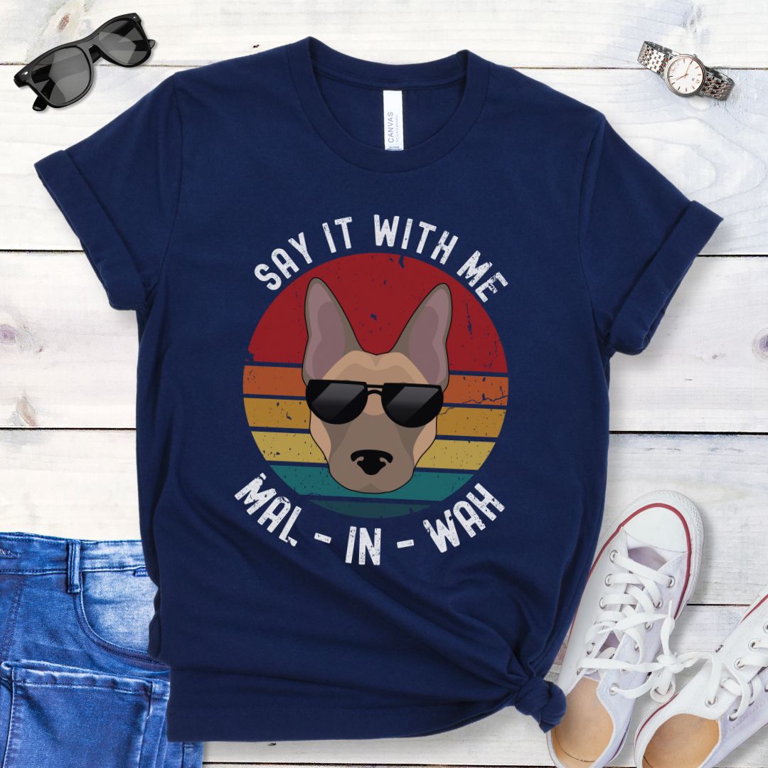 Navy t-shirt for Belgian Malinois lovers with a Malinois dog face wearing sunglasses, on a vintage sunset graphic, and humorous text 'Say it with me - Mal-in-wah', shown on a wooden surface with accessories.