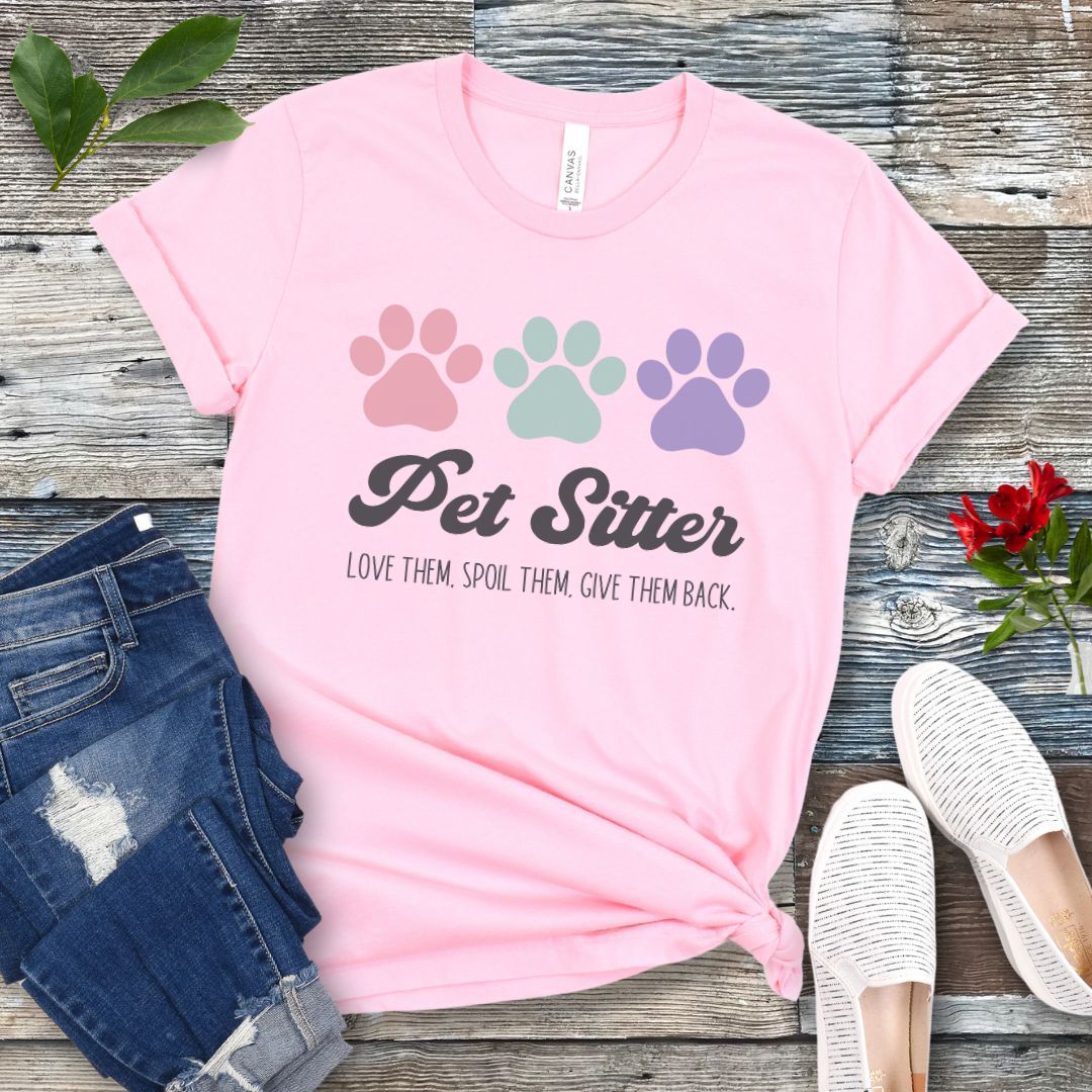 A pink unisex short-sleeve T-shirt with 'Pet Sitter: Love Them, Spoil Them, Give Them Back' text and colorful paw prints design, laid flat with jeans and white shoes on a wooden surface.