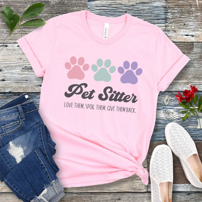 A pink unisex short-sleeve T-shirt with 'Pet Sitter: Love Them, Spoil Them, Give Them Back' text and colorful paw prints design, laid flat with jeans and white shoes on a wooden surface.