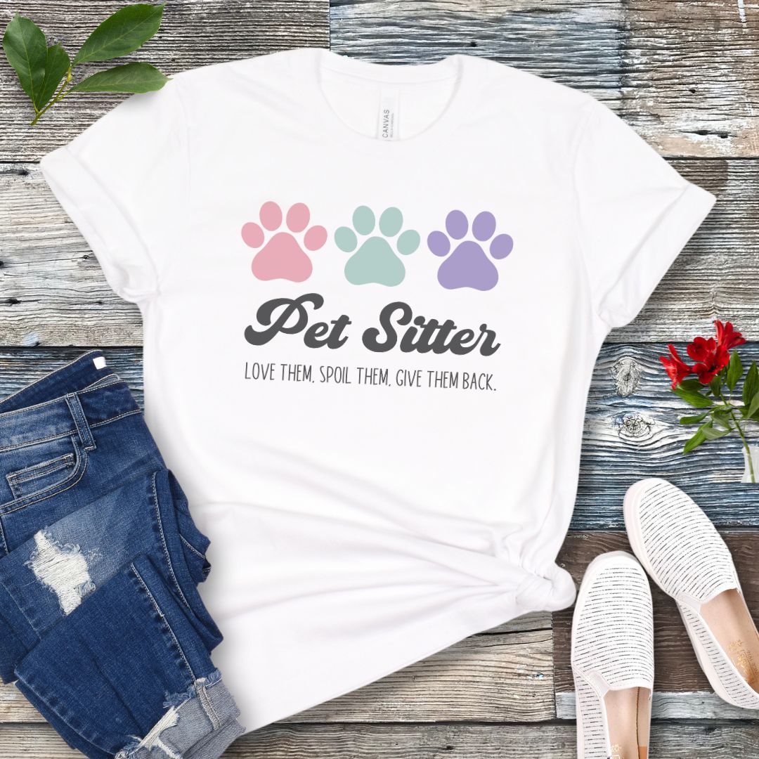 A white unisex short-sleeve T-shirt with 'Pet Sitter: Love Them, Spoil Them, Give Them Back' text and colorful paw prints design, laid flat with jeans and white shoes on a wooden surface.
