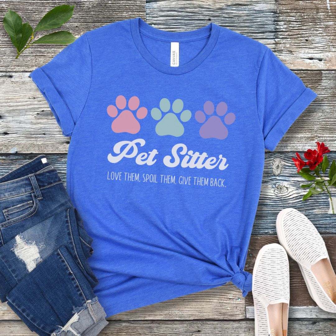A royal blue unisex short-sleeve T-shirt with 'Pet Sitter: Love Them, Spoil Them, Give Them Back' text and colorful paw prints design, laid flat with jeans and white shoes on a wooden surface.
