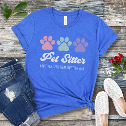 A royal blue unisex short-sleeve T-shirt with 'Pet Sitter: Love Them, Spoil Them, Give Them Back' text and colorful paw prints design, laid flat with jeans and white shoes on a wooden surface.
