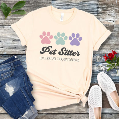 A beige unisex short-sleeve T-shirt with 'Pet Sitter: Love Them, Spoil Them, Give Them Back' text and colorful paw prints design, laid flat with jeans and white shoes on a wooden surface.