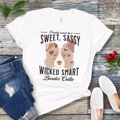 White t-shirt for Border Collie lovers with a graphic of a Red Merle Border Collie face and the humorous text, 'Proudly Owned By a Sweet, Sassy, Wicked Smart Border Collie' on a wooden surface with jeans and shoes.
