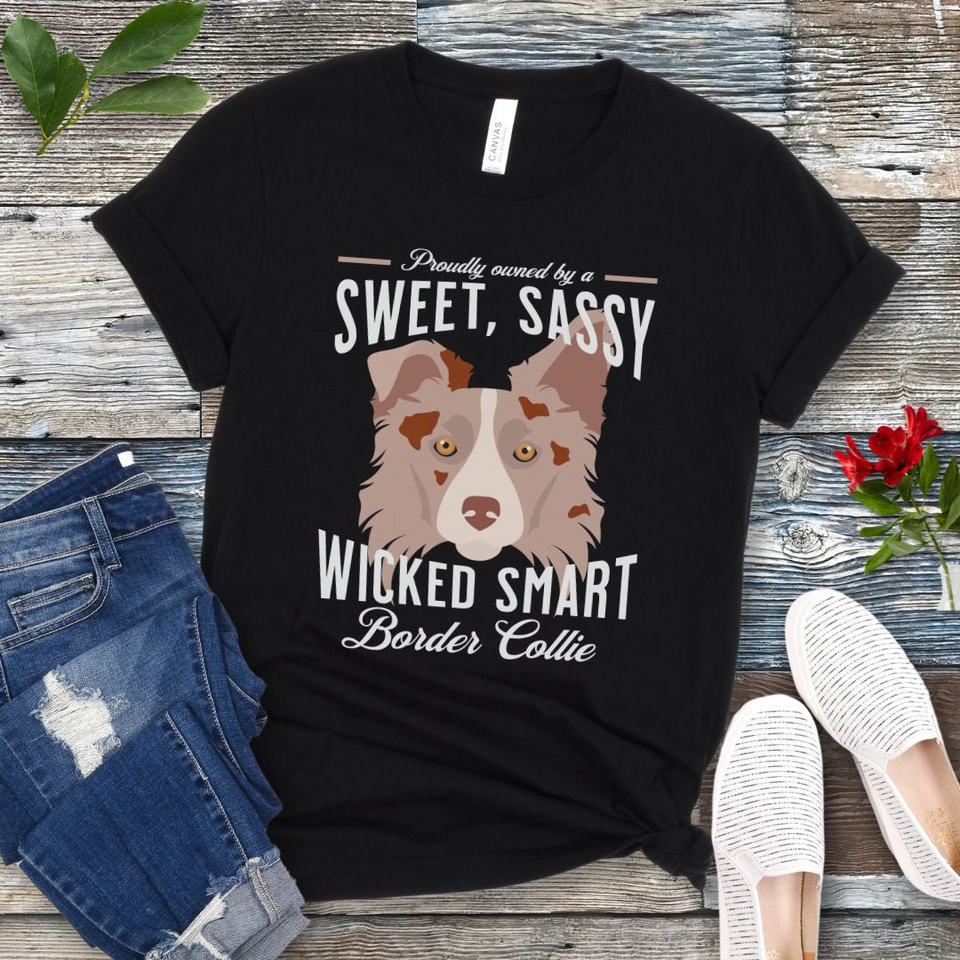 Black t-shirt for Border Collie lovers with a graphic of a Red Merle Border Collie face and the humorous text, 'Proudly Owned By a Sweet, Sassy, Wicked Smart Border Collie' on a wooden surface with jeans and shoes.