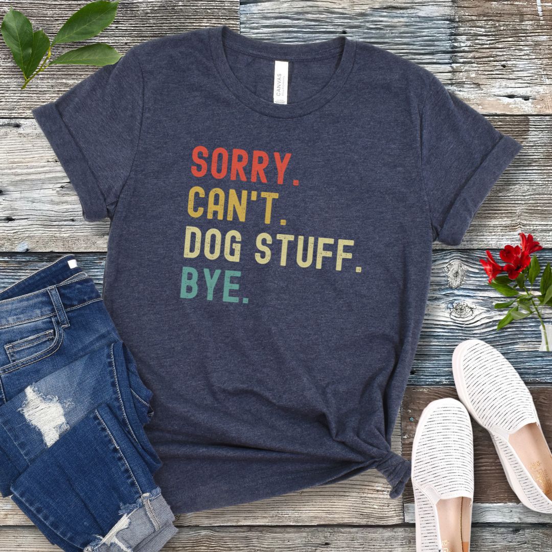 A heather navy blue unisex short sleeve T-shirt with colorful stacked text 'SORRY. CAN'T. DOG STUFF. BYE.', presented on a wooden surface with jeans and shoes.
