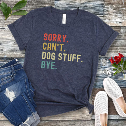 A heather navy blue unisex short sleeve T-shirt with colorful stacked text 'SORRY. CAN'T. DOG STUFF. BYE.', presented on a wooden surface with jeans and shoes.