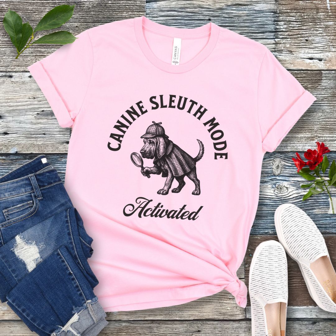 Pink scent detection t-shirt with a vintage style graphic of a bloodhound in Sherlock Holmes' apparel, and 'Canine Sleuth Mode - Activated' text , presented on a wooden surface with jeans and shoes.