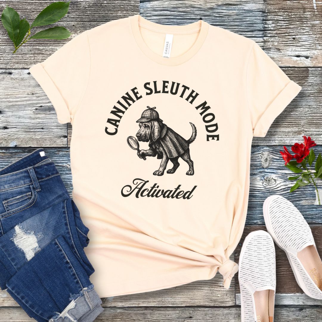 Beige scent detection t-shirt with a vintage style graphic of a bloodhound in Sherlock Holmes' apparel, and 'Canine Sleuth Mode - Activated' text , presented on a wooden surface with jeans and shoes.