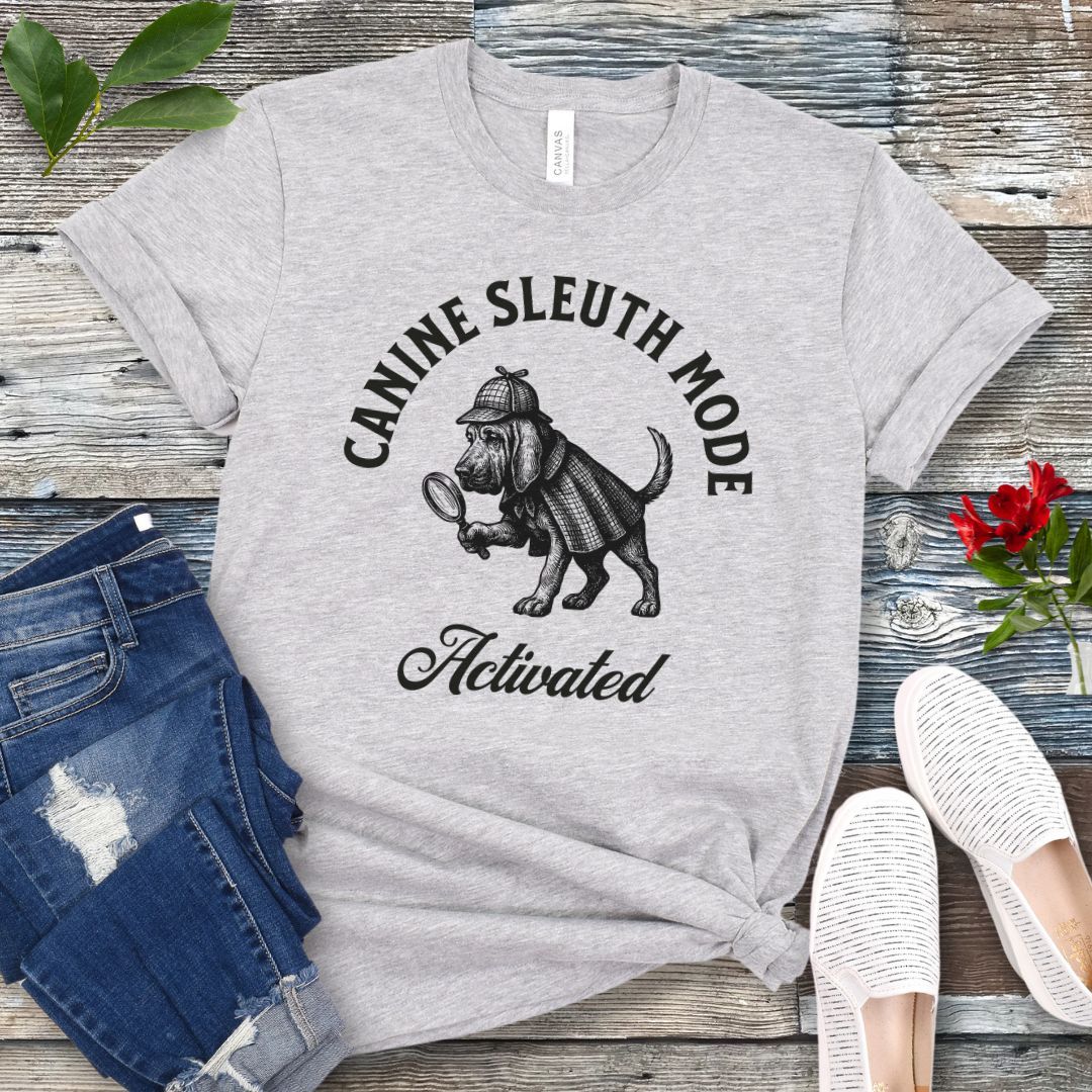Grey scent detection t-shirt with a vintage style graphic of a bloodhound in Sherlock Holmes' apparel, and 'Canine Sleuth Mode - Activated' text , presented on a wooden surface with jeans and shoes.