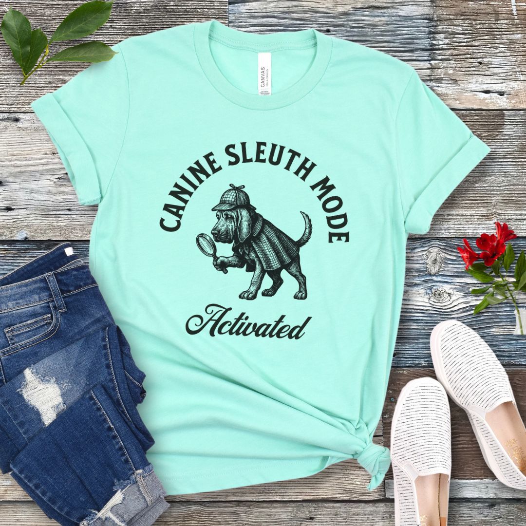 Mint green scent detection t-shirt with a vintage style graphic of a bloodhound in Sherlock Holmes' apparel, and 'Canine Sleuth Mode - Activated' text , presented on a wooden surface with jeans and shoes.