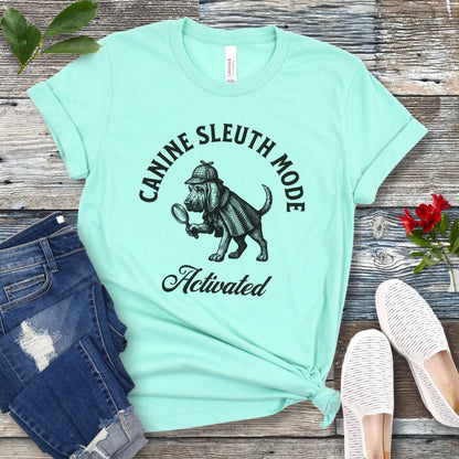 Mint green scent detection t-shirt with a vintage style graphic of a bloodhound in Sherlock Holmes' apparel, and 'Canine Sleuth Mode - Activated' text , presented on a wooden surface with jeans and shoes.