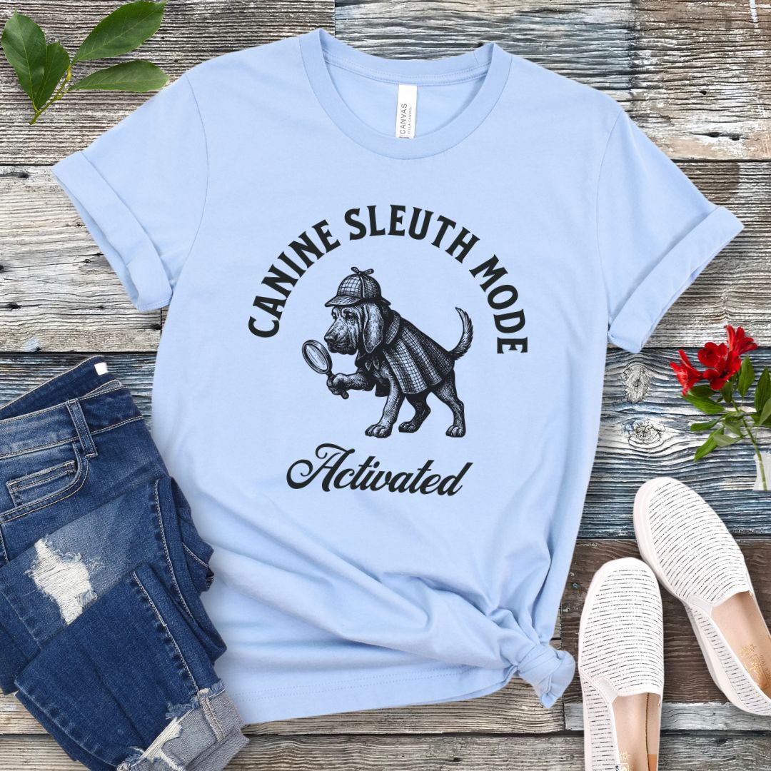 Light blue scent detection t-shirt with a vintage style graphic of a bloodhound in Sherlock Holmes' apparel, and 'Canine Sleuth Mode - Activated' text , presented on a wooden surface with jeans and shoes.