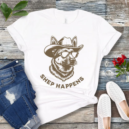 White t-shirt with a graphic of a German Shepherd dog wearing sunglasses and a cowboy hat, with text 'SHEP HAPPENS', displayed on a wooden surface with jeans and shoes.