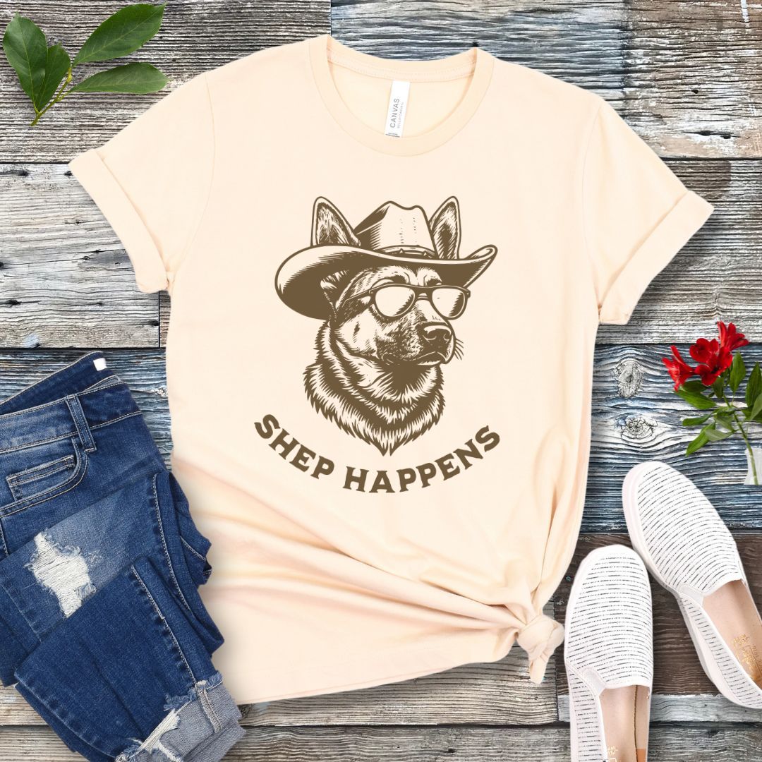 Beige t-shirt with a graphic of a German Shepherd dog wearing sunglasses and a cowboy hat, with text 'SHEP HAPPENS', displayed on a wooden surface with jeans and shoes.