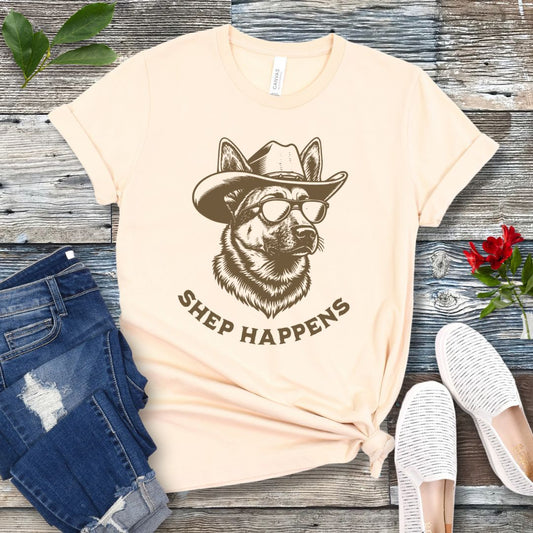 Beige t-shirt with a graphic of a German Shepherd dog wearing sunglasses and a cowboy hat, with text 'SHEP HAPPENS', displayed on a wooden surface with jeans and shoes.