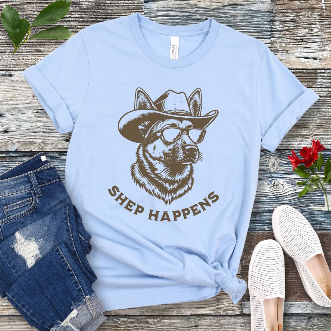 Light blue t-shirt with a graphic of a German Shepherd dog wearing sunglasses and a cowboy hat, with text 'SHEP HAPPENS', displayed on a wooden surface with jeans and shoes.