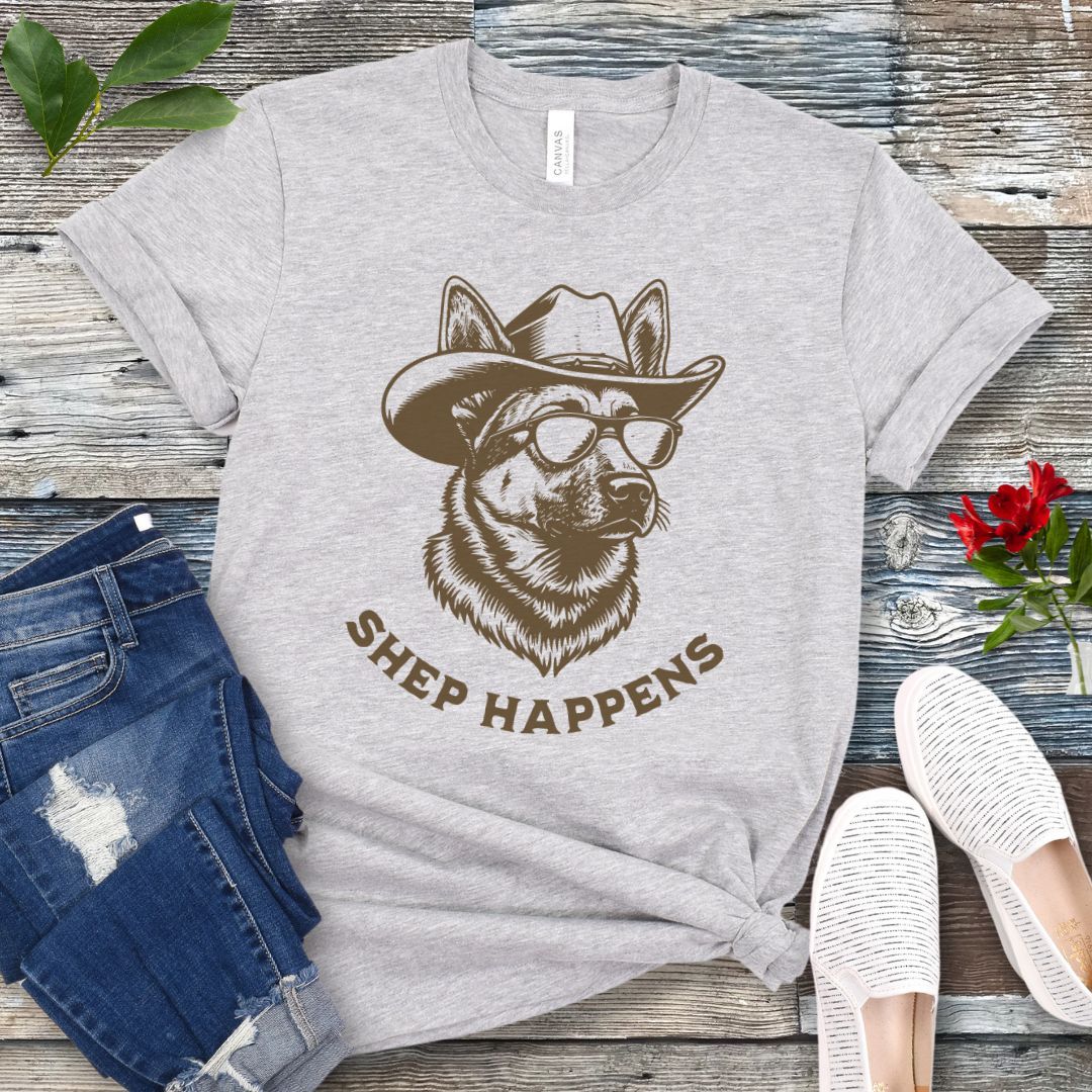 Grey t-shirt with a graphic of a German Shepherd dog wearing sunglasses and a cowboy hat, with text 'SHEP HAPPENS', displayed on a wooden surface with jeans and shoes.