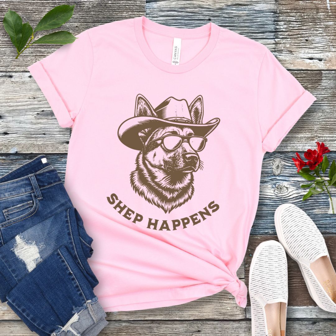 Pink t-shirt with a graphic of a German Shepherd dog wearing sunglasses and a cowboy hat, with text 'SHEP HAPPENS', displayed on a wooden surface with jeans and shoes.