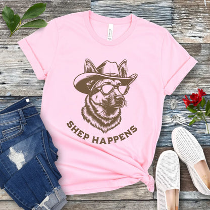 Pink t-shirt with a graphic of a German Shepherd dog wearing sunglasses and a cowboy hat, with text 'SHEP HAPPENS', displayed on a wooden surface with jeans and shoes.